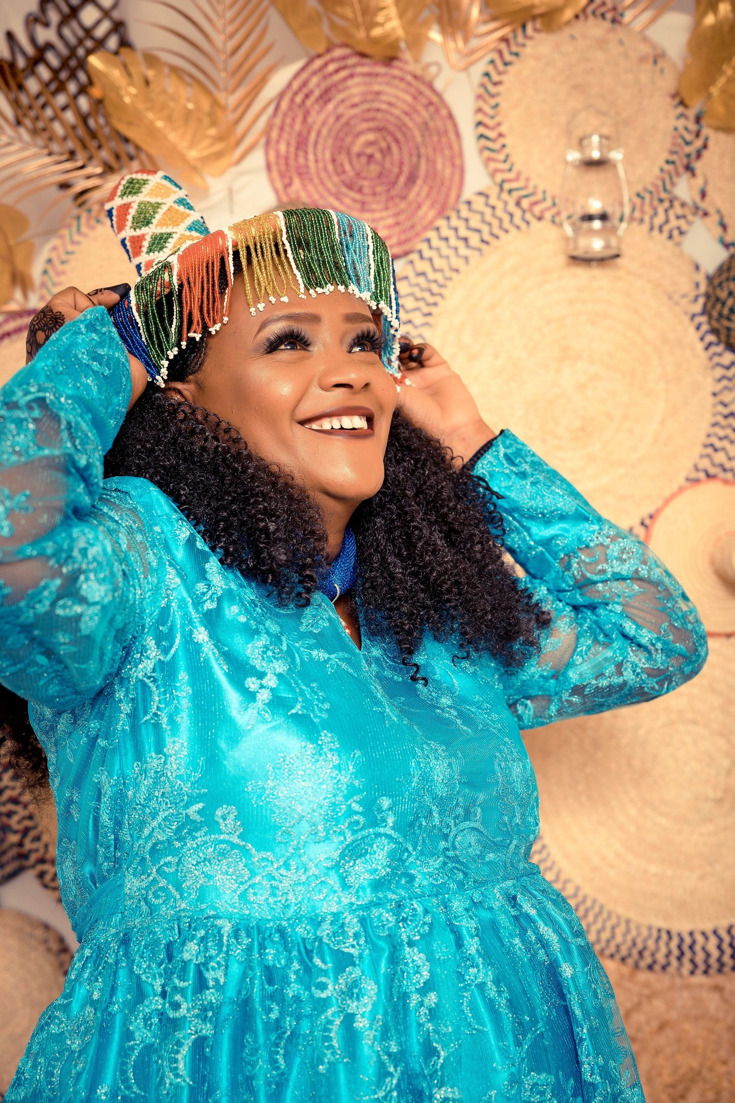 Woman in turquoise lace dress and headpiece smiling against decorative background.