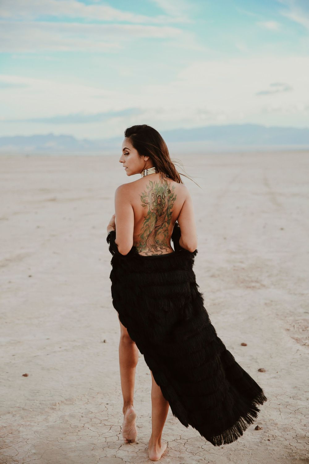 Barefoot woman with intricate back tattoo drapes black shawl while standing on sandy desert landscape.
