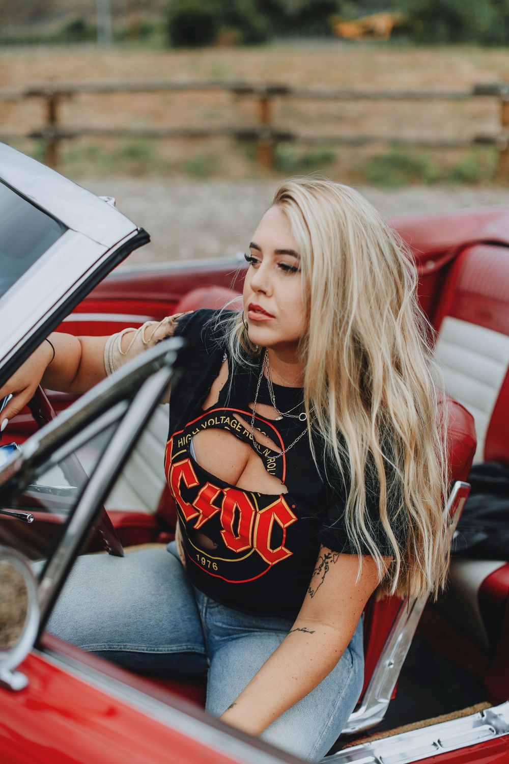 Classic rock fan in AC/DC t-shirt and jeans poses in vintage red convertible car at sunset.