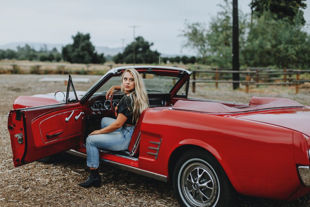 A classic red Ford Mustang convertible parked on a gravel lot with the driver's door open.