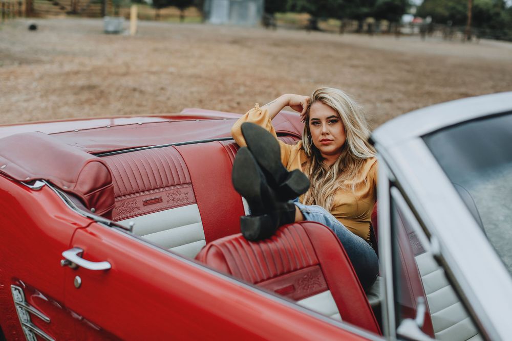 A woman with blonde hair sits in a vintage red convertible car with leather interior during golden hour lighting.