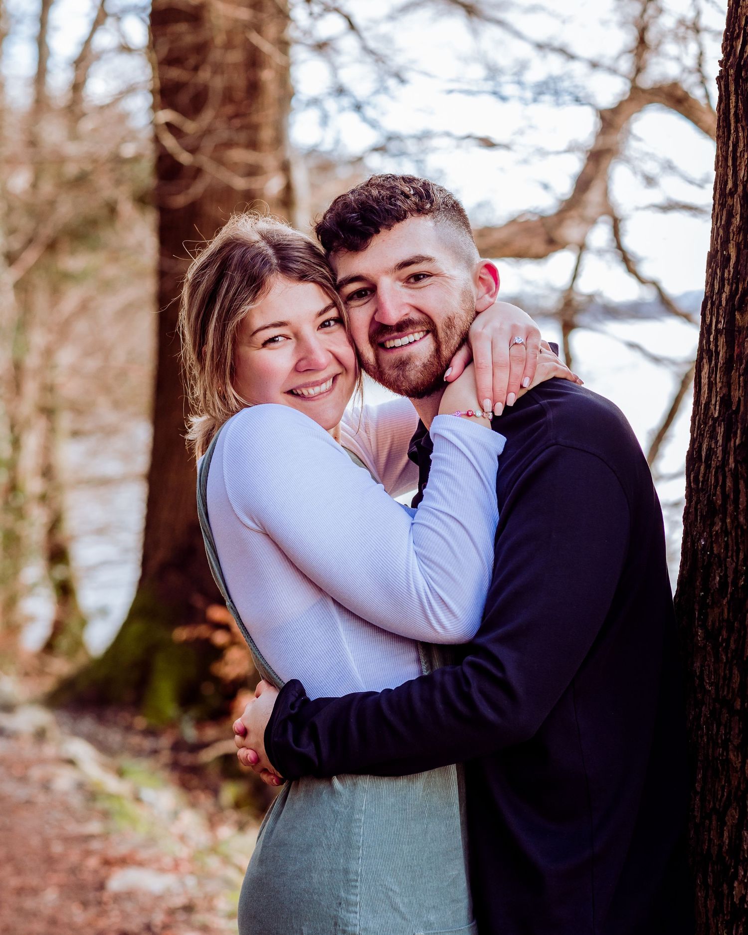 Couple embracing among trees on a forest trail near water.