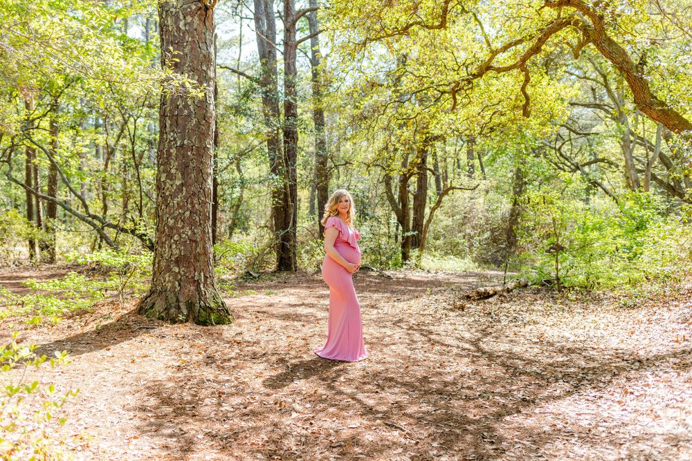 Pregnant woman in flowing pink dress stands alone in dappled sunlight of wooded forest.