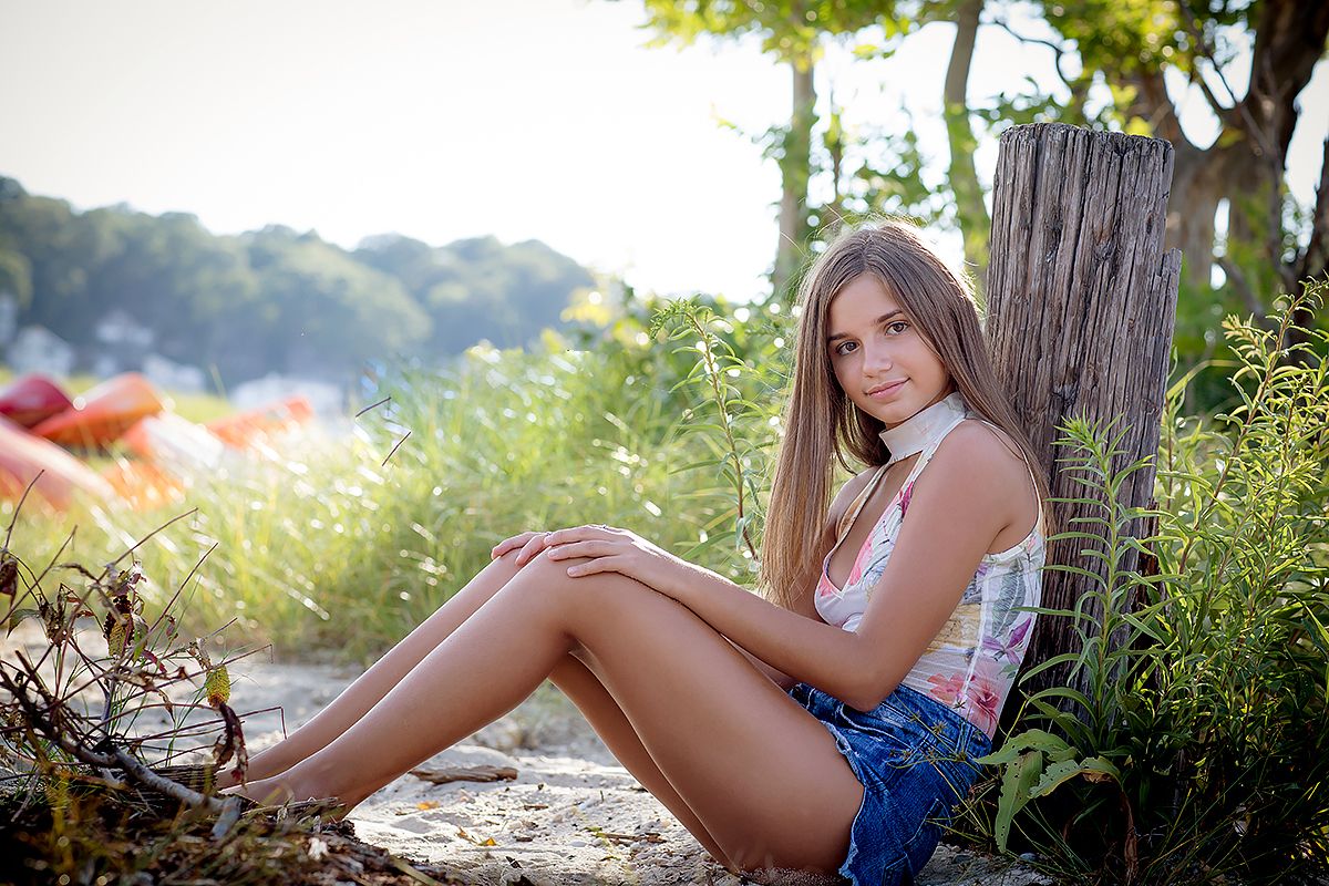 Person in white top and denim shorts sitting by wooden post near water.