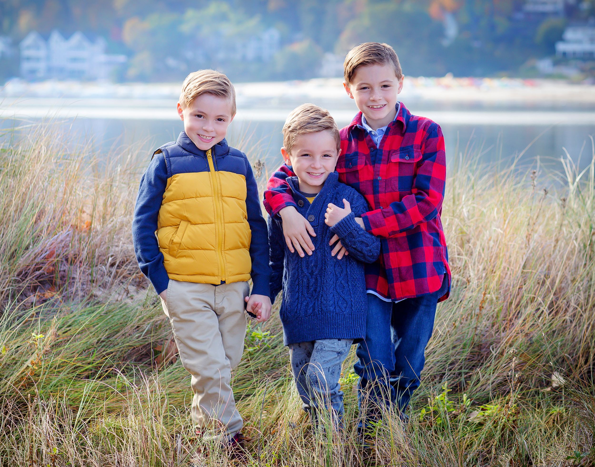 Three young boys in casual outerwear pose together in tall grass by lakeside.