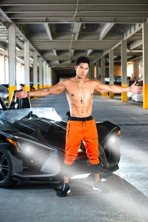 Muscular figure in orange pants strikes pose next to luxury car in covered garage.