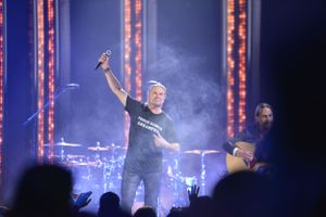 Concert performer raising arm triumphantly on stage with dramatic lighting and smoke effects in background.
