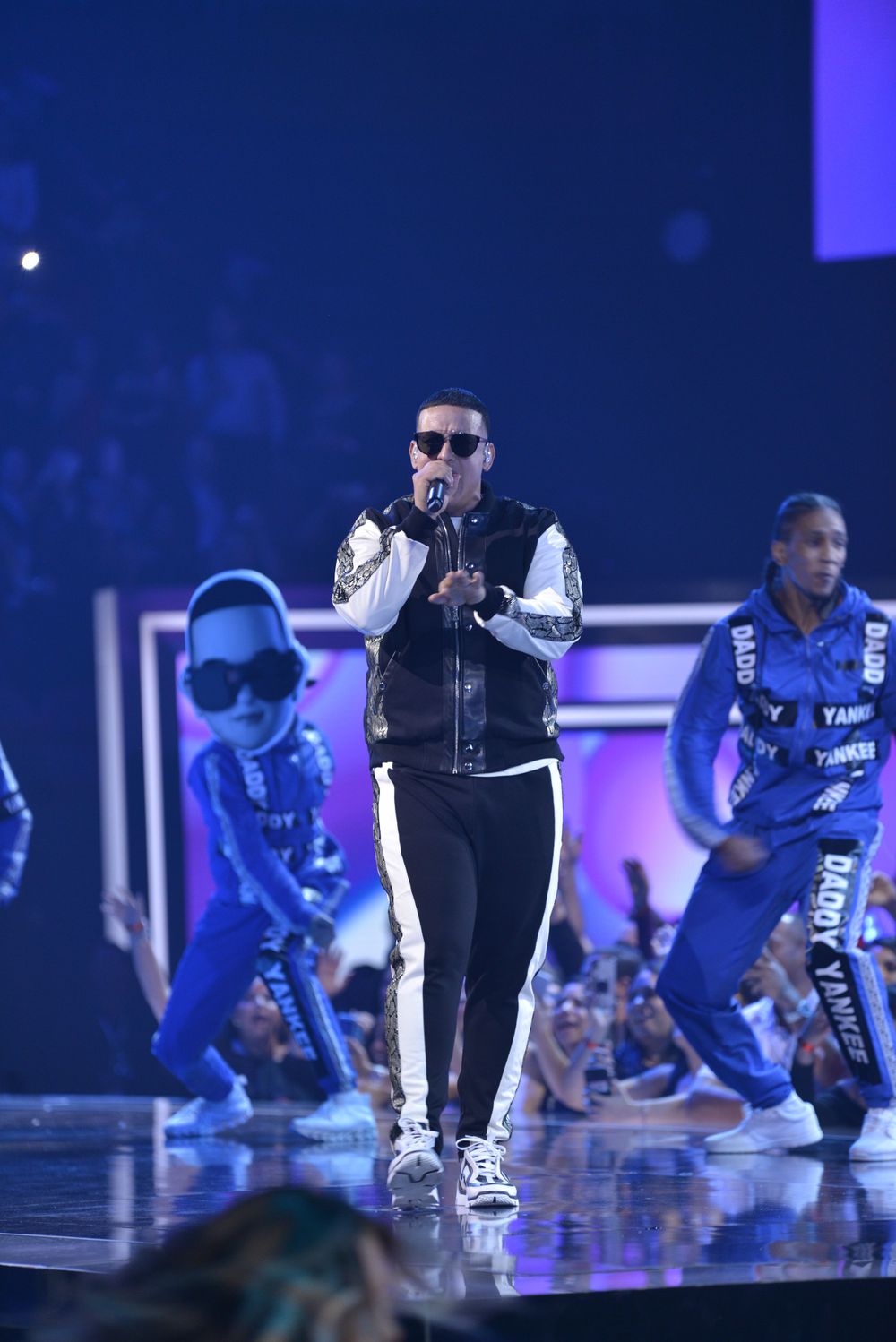 Concert performer in black vest and sunglasses with dancers in blue costumes on stage.