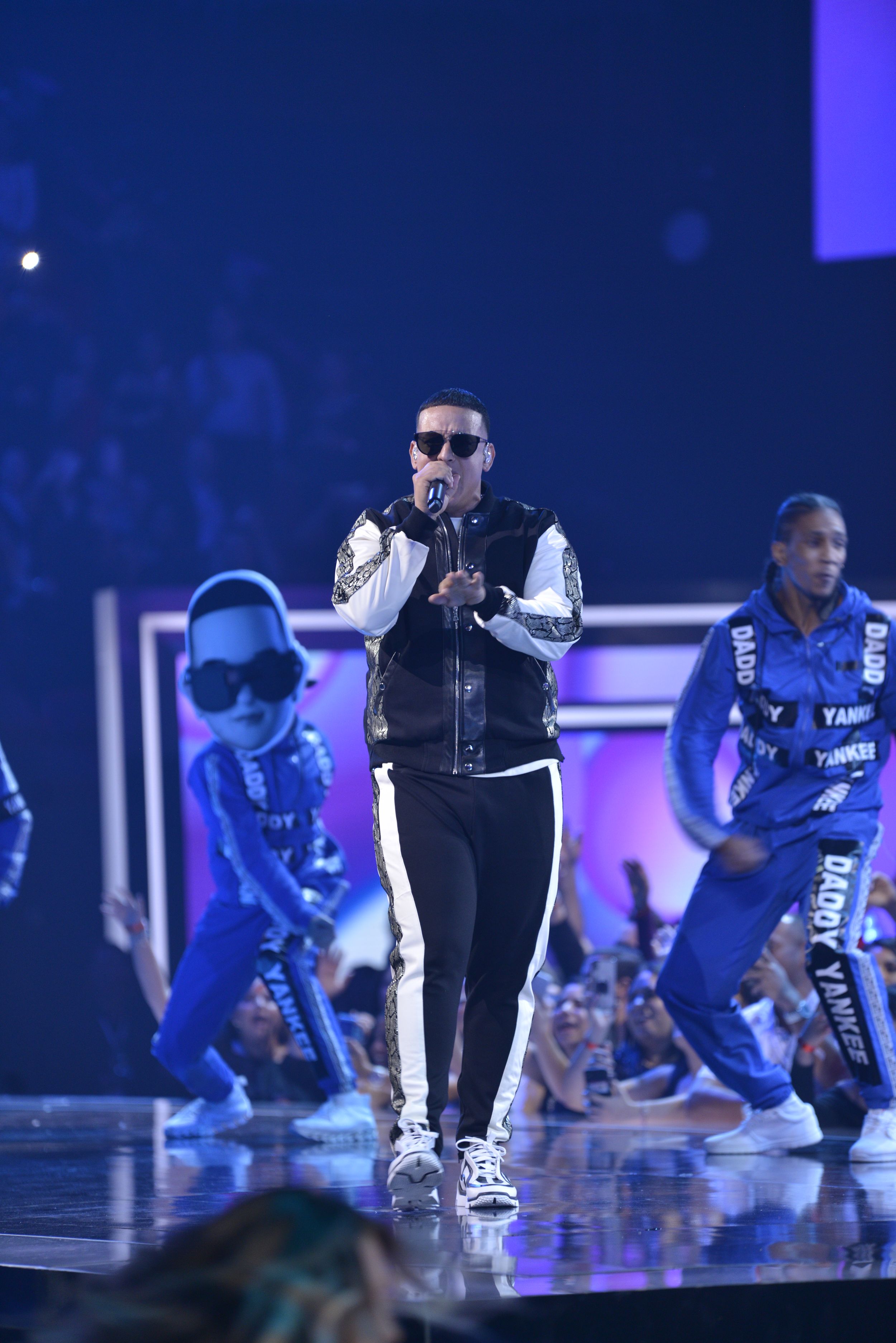 Concert performer in black vest and sunglasses with dancers in blue costumes on stage.