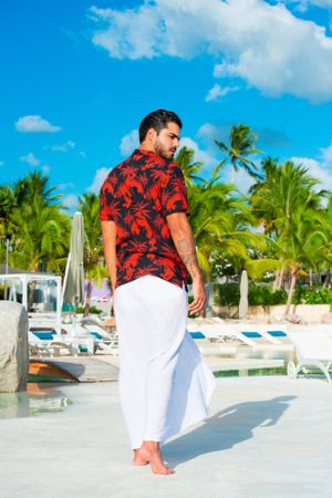 Person wearing red floral shirt and white pants on tropical beach.