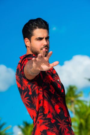 Person in red floral shirt making dramatic hand gesture against tropical background.