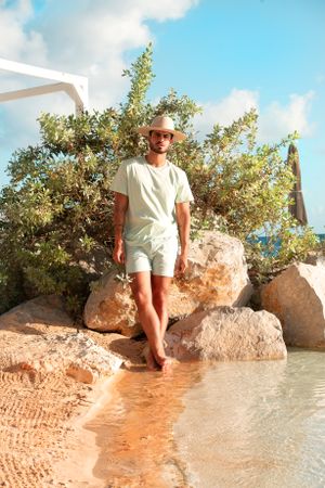 Person in all-white summer outfit standing on rocky beach with palm trees.