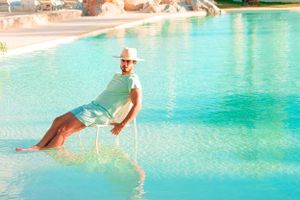 Person relaxing in chair by turquoise pool wearing beach attire.