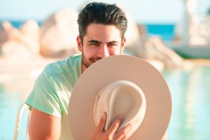 Person holding beige sun hat near tropical pool setting.