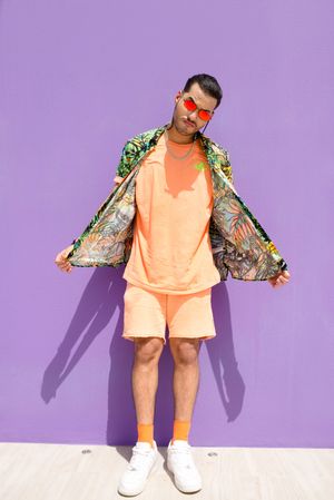 Stylish man in orange shorts and floral jacket poses against purple wall.