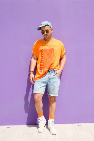 Man in orange t-shirt and denim shorts poses casually against purple wall.