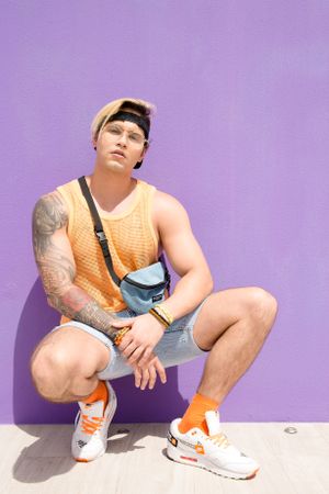 Athletic person in orange tank top and denim shorts squats against purple wall.