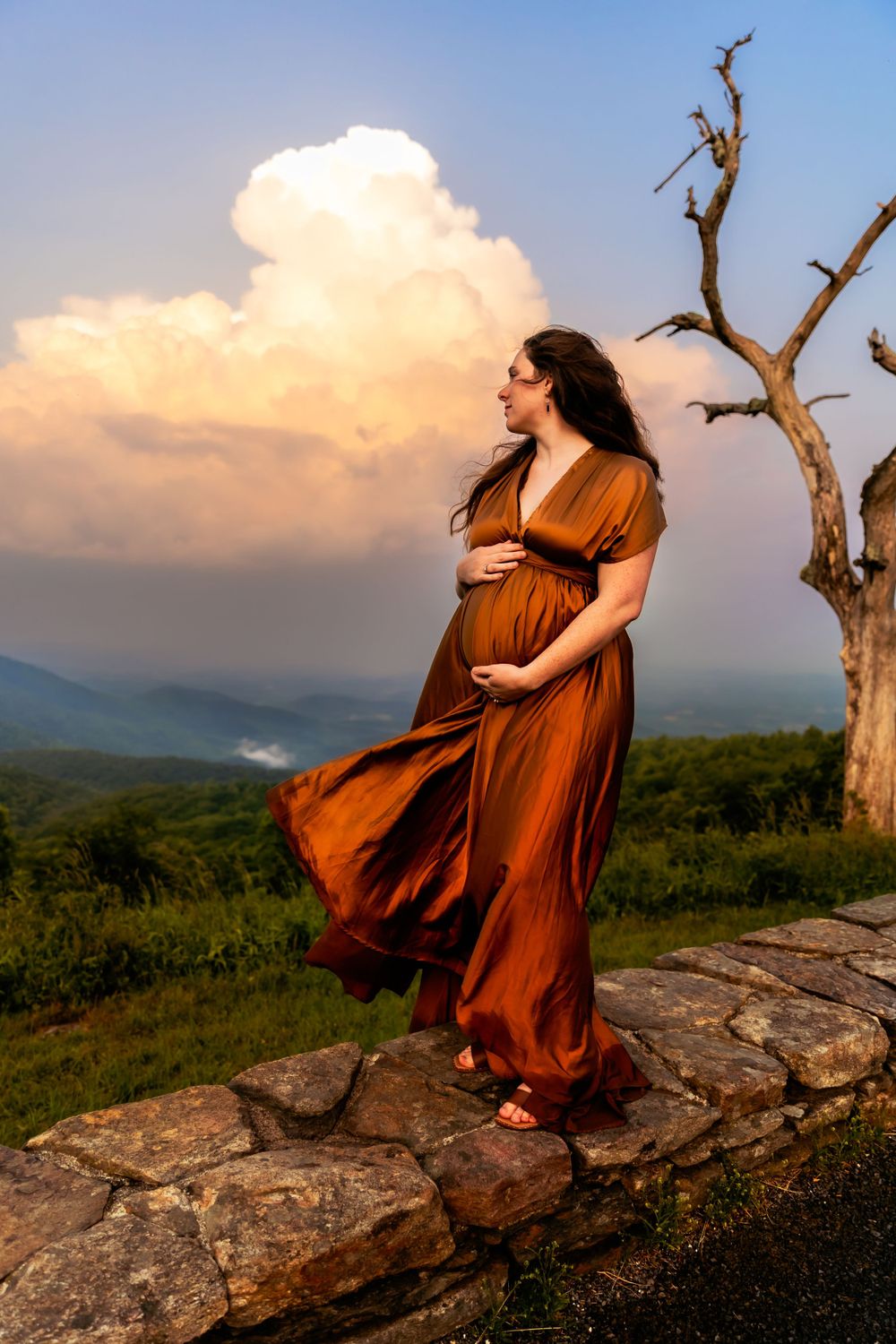 Pregnant woman in rust gown poses on rocky overlook with dramatic clouds at sunset.