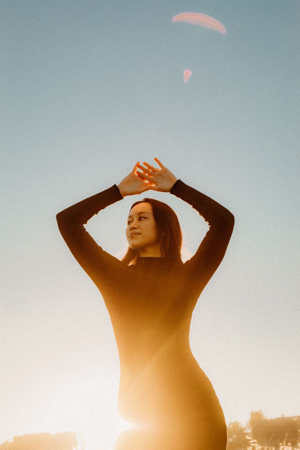 Silhouetted figure of model stretches arms up at sunset with lens sun flares in the blue sky above in Anaheim California