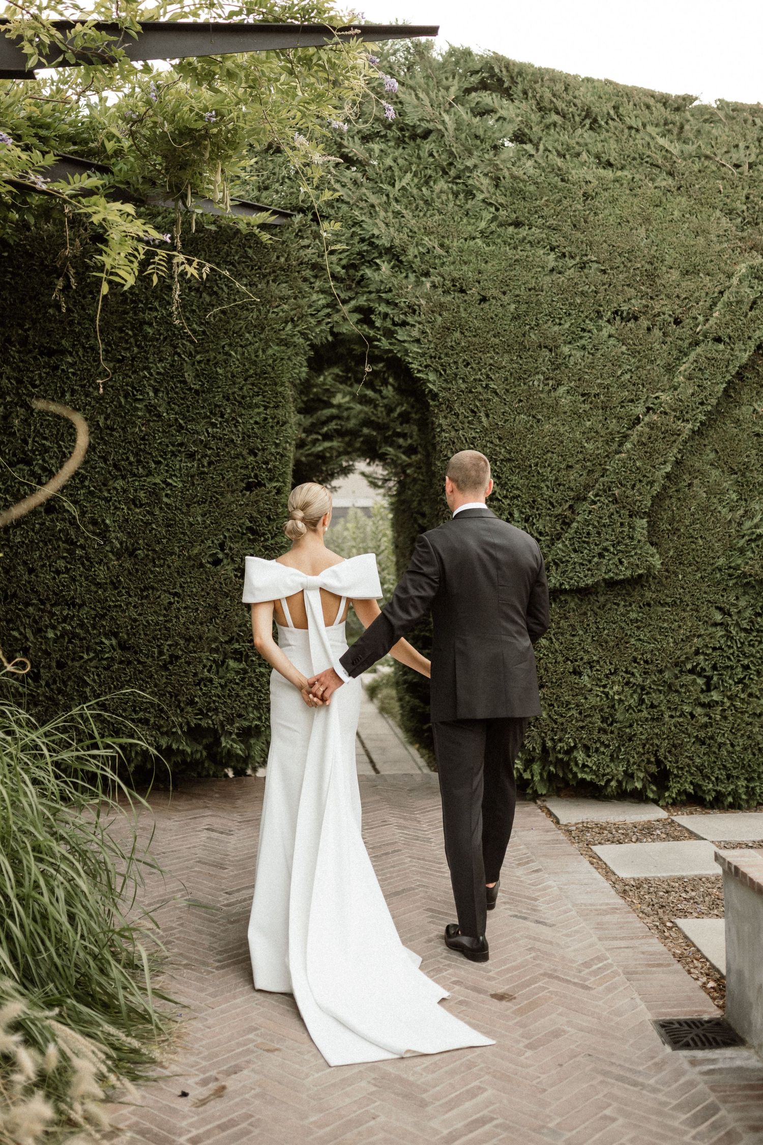 Bride and groom with their arms intertwined, walking away from the camera towards a lush, green hedge.
