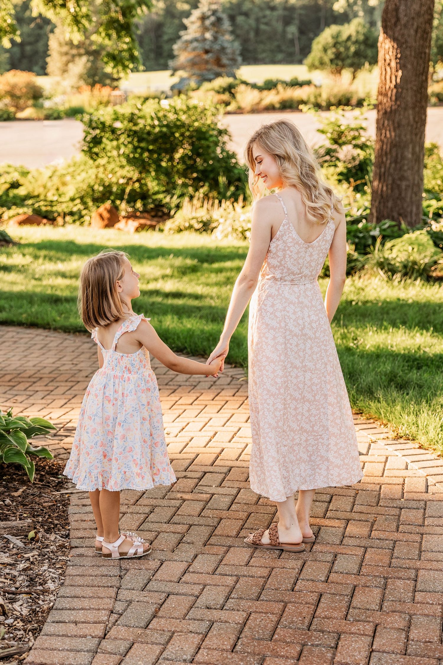 Two people in floral dresses hold hands while walking on brick path in garden.