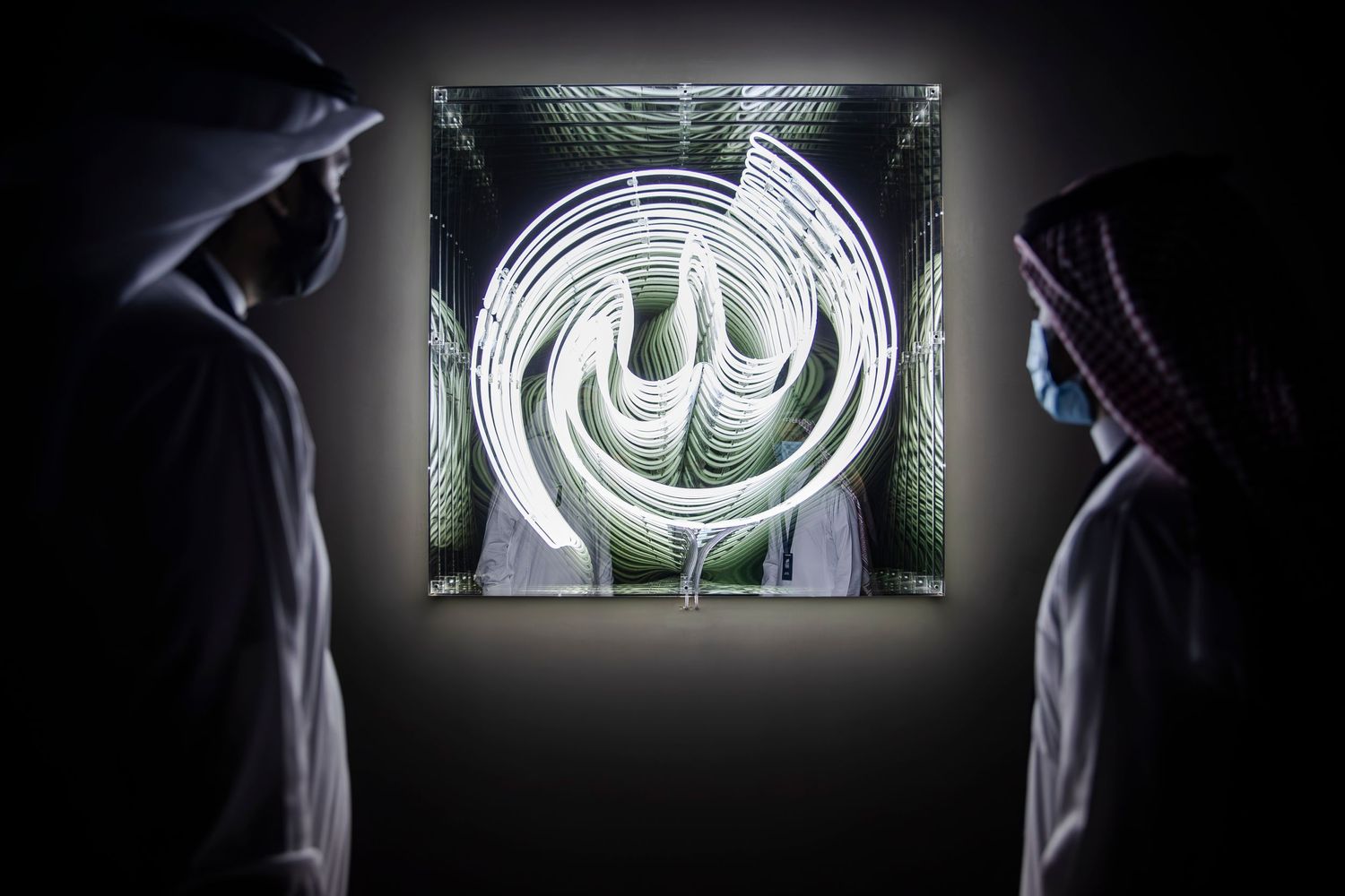 Two silhouettes view a glowing white Arabic calligraphy art installation in a dark space.