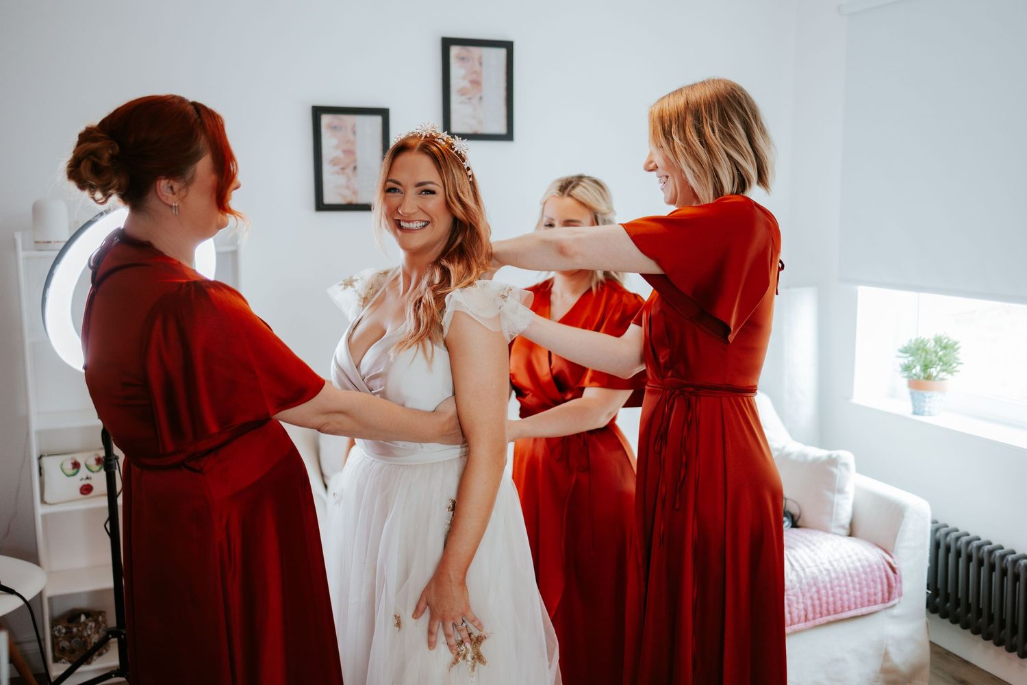 Bridesmaids in red dresses help a bride get ready on her wedding day in a bright room.
