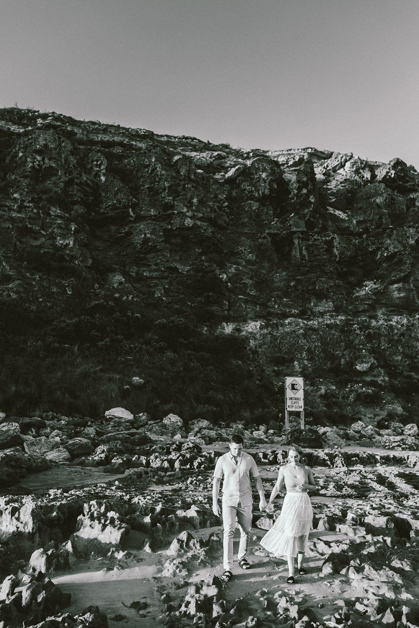 A couple walks together on dark rocky terrain with dramatic cliffs in the background.