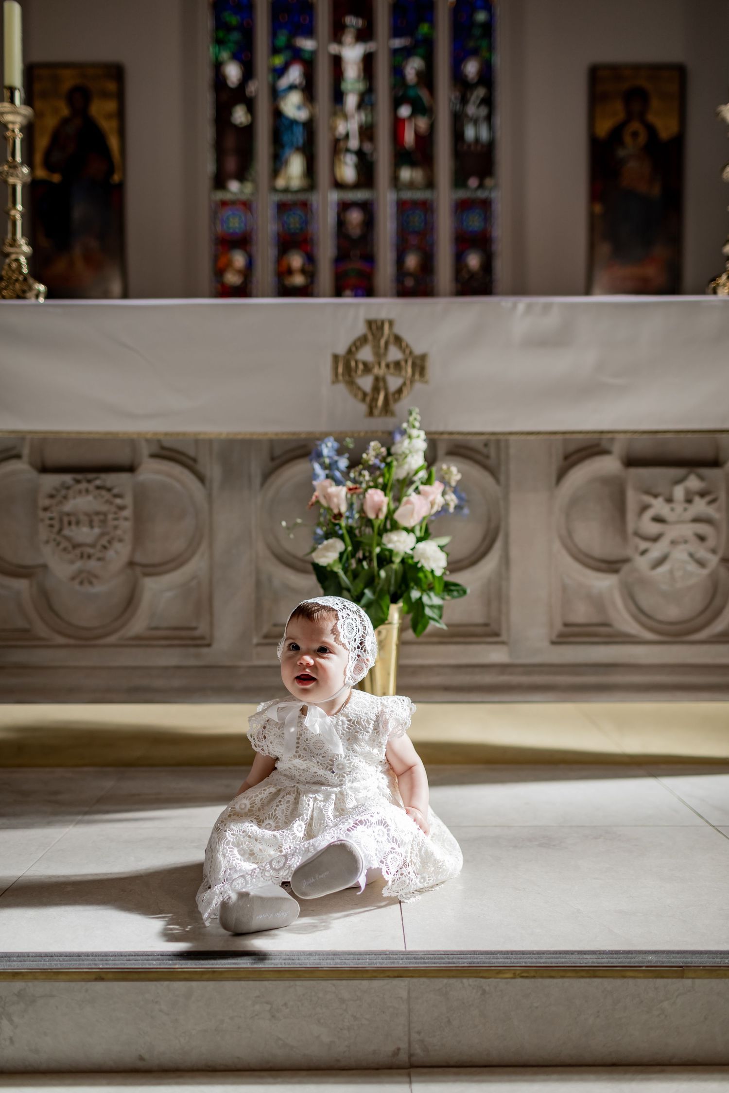 A small child in white baptismal dress sits near stained glass windows and floral arrangements in church.