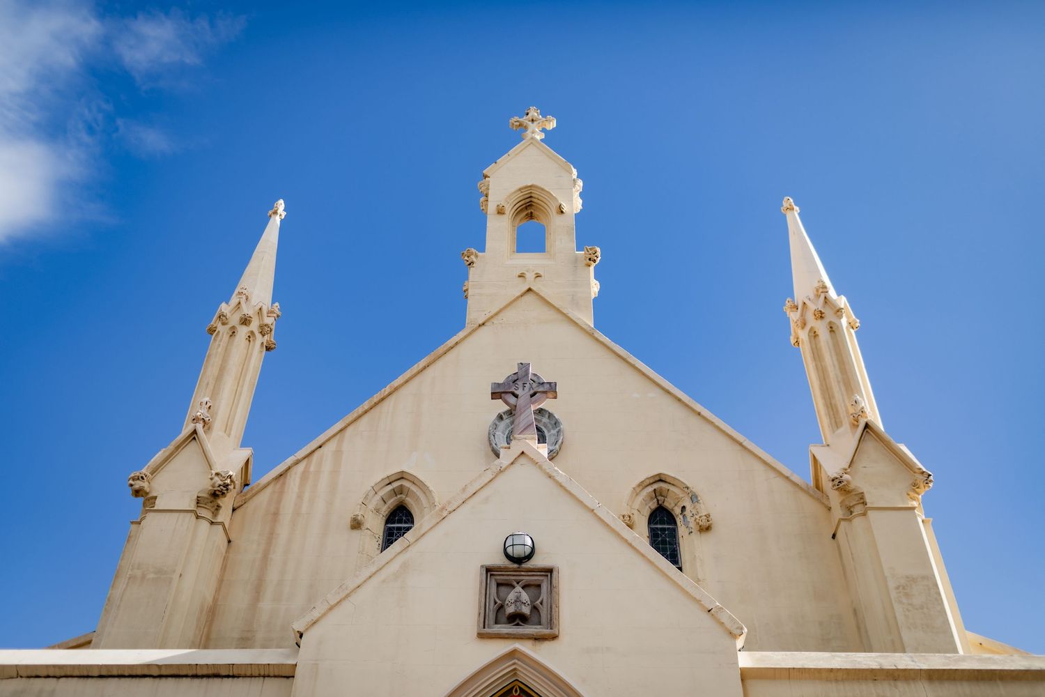 White gothic church facade with ornate spires and cross against bright blue sky.