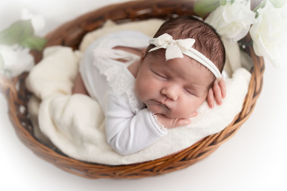 Newborn baby girl cuddled in a basket wearing a white outfit and white bow headband.