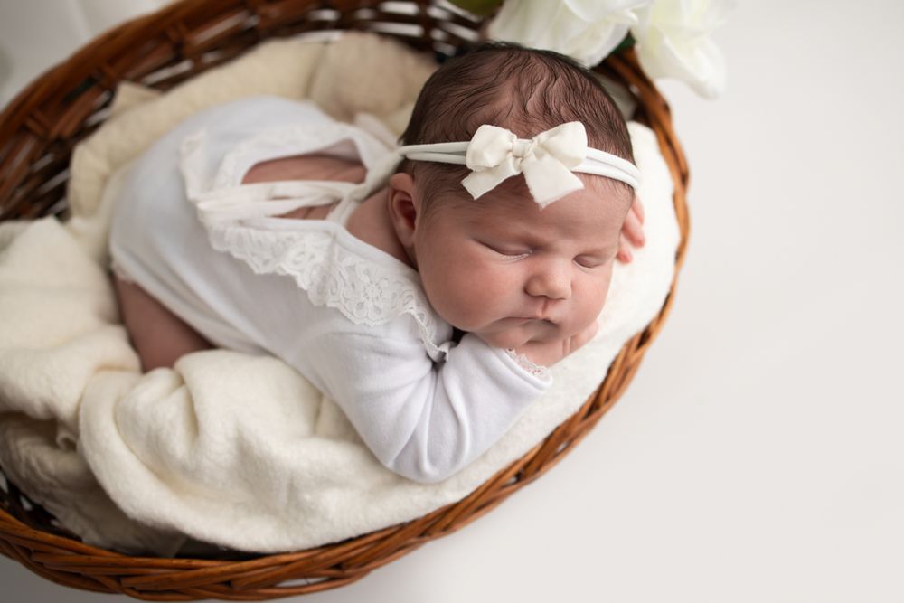 Newborn baby girl posed on her belly in a basket wearing a white outfit and bow headband.