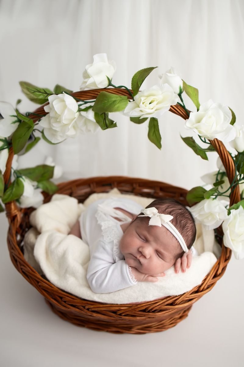 Newborn baby girl posed in a brown woven basket with flowers of the basket handle.  She is wearing a white outfit.