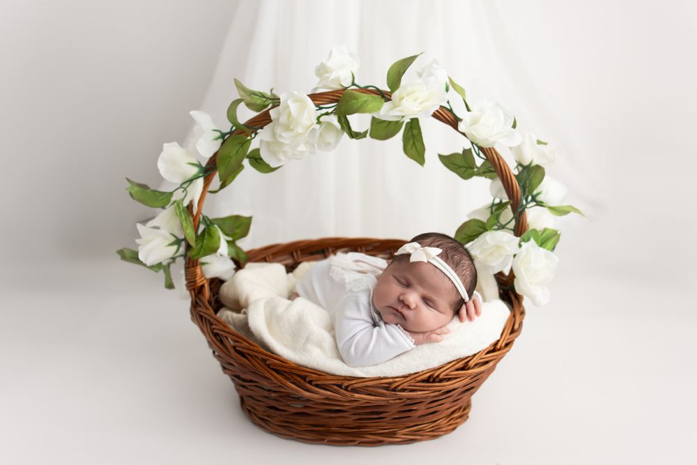 Newborn baby girl posed in a basket with flowers hanging over the handle and flowing curtains in the background.