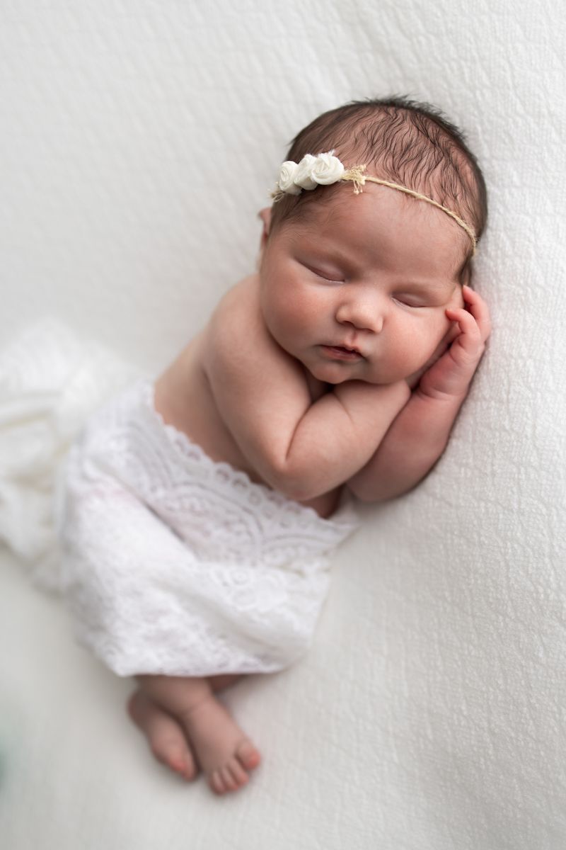 Newborn baby girl laying on her side with a white wrap over her legs.  Both hands are under her cheek.