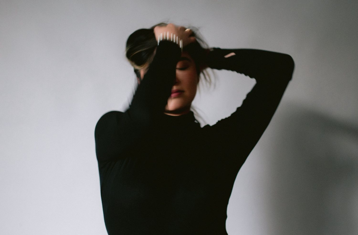 Artistic black and white portrait of someone in a dark turtleneck sweater with hands raised against a light background.