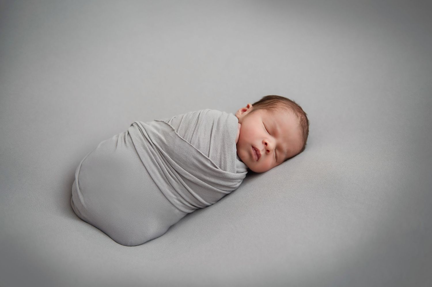 Newborn baby sleeping peacefully while wrapped in light grey swaddle.