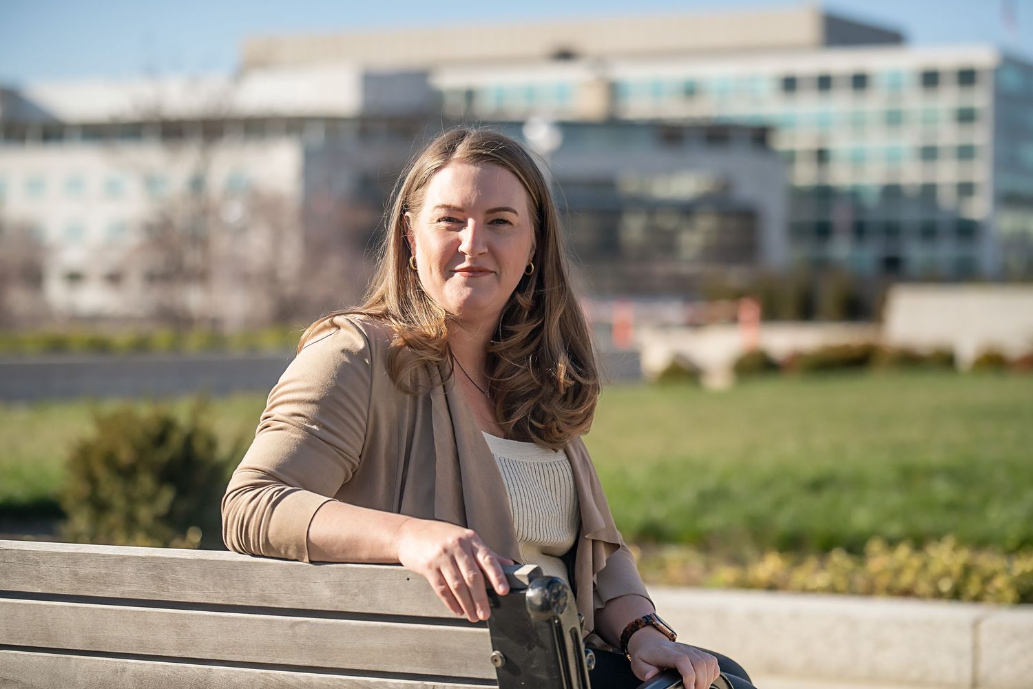 A person sits on a bench outdoors with urban buildings visible in the background.