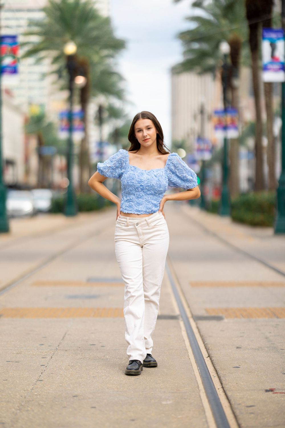 Woman walks down palm-lined street wearing blue puff sleeve top and white pants.