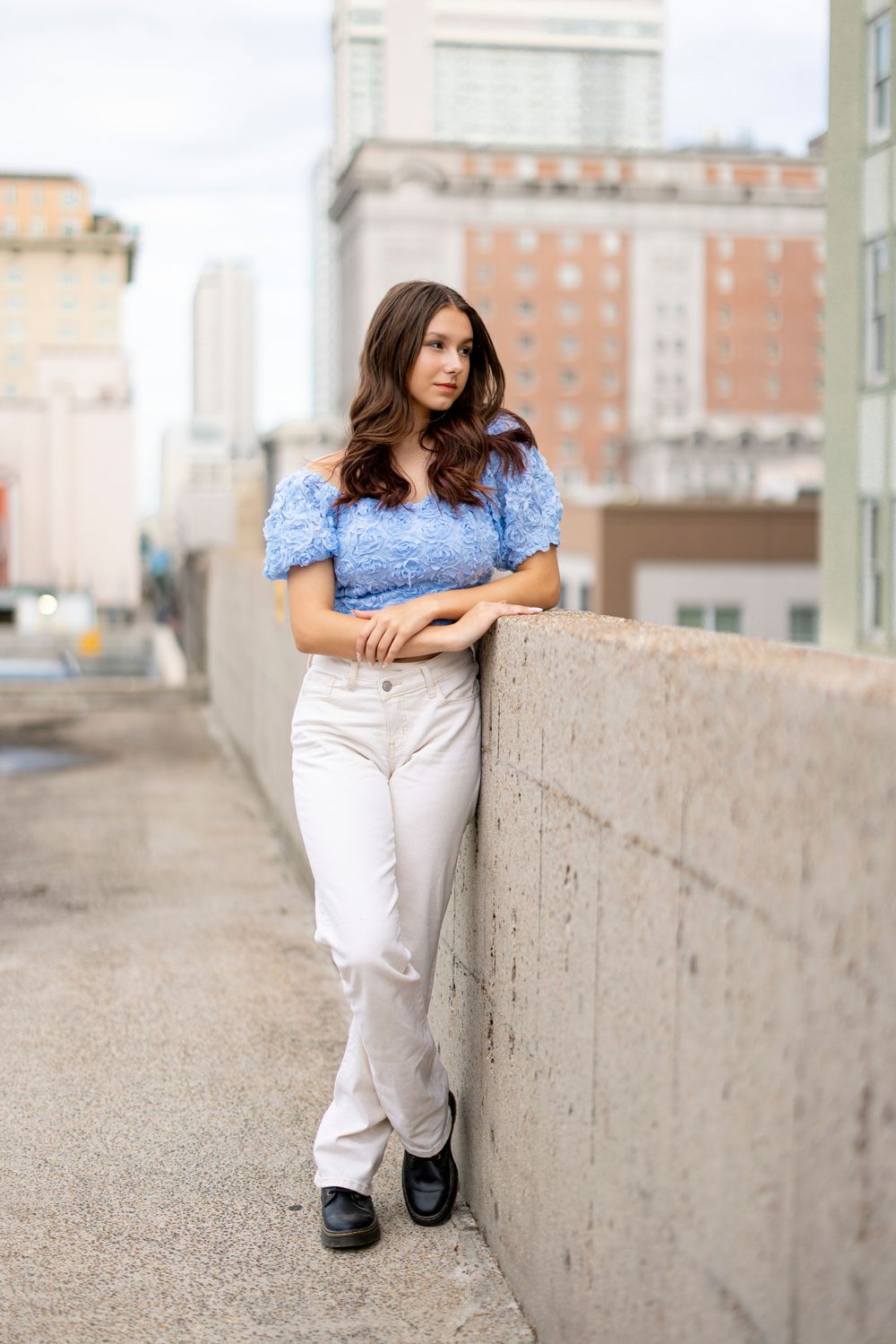 Woman in blue top and white pants leans against concrete wall in urban setting.