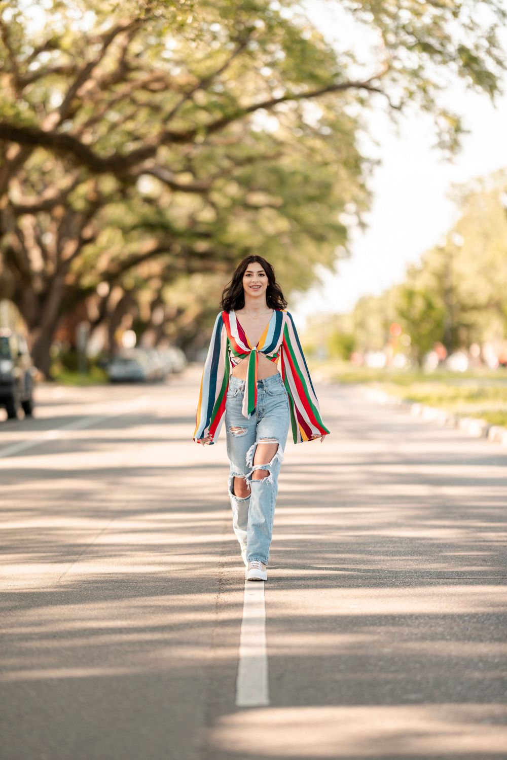 A casual street style outfit featuring distressed jeans and colorful striped cardigan on a tree-lined path.