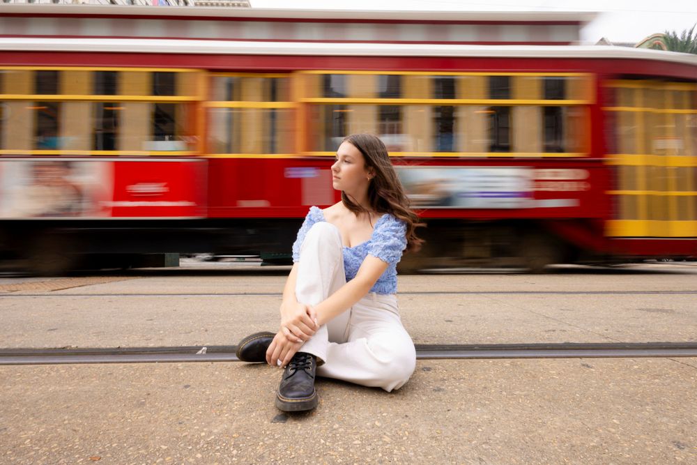 A person sits casually on the ground as a red and yellow train car blurs past in motion.