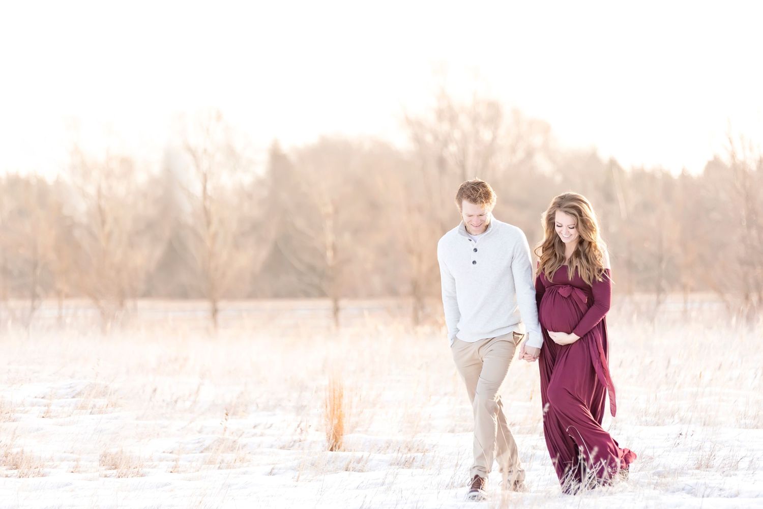 Couple walking hand in hand through snowy field at sunset wearing white and burgundy outfits.