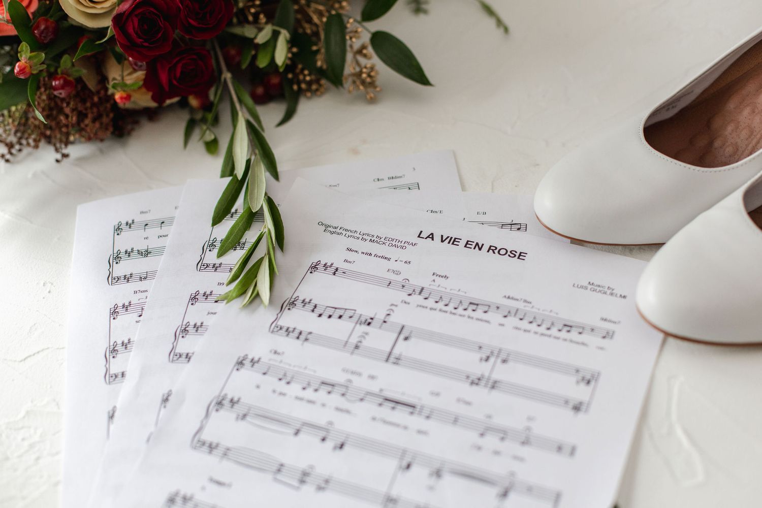 Sheet music and white bridal shoes displayed with burgundy floral arrangement.