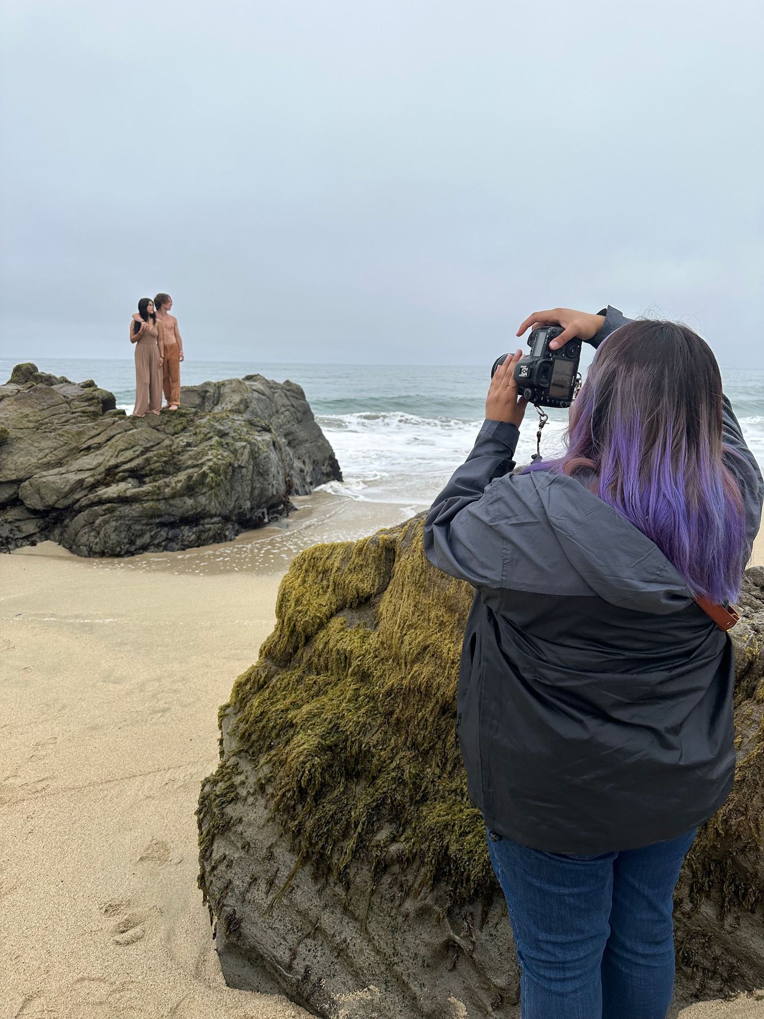 Photographer with purple hair taking pictures of subjects on rocky beach cliff.
