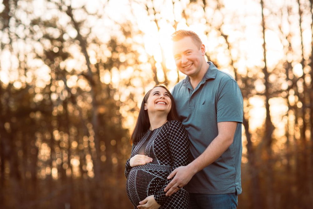 A couple shares an intimate moment at sunset in the woods while expecting their first child.