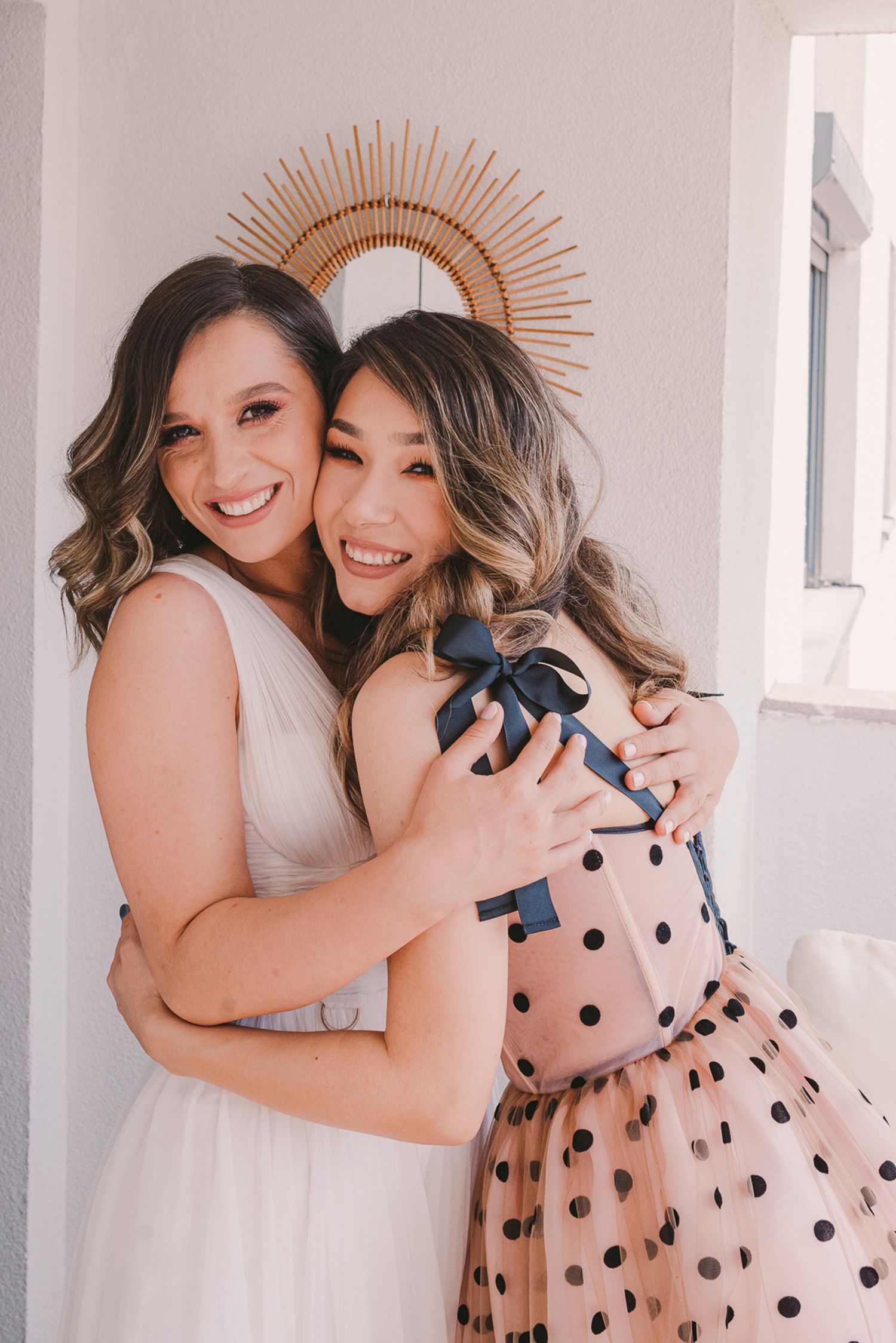 Two friends share a joyful embrace while wearing polka dot and white dresses.
