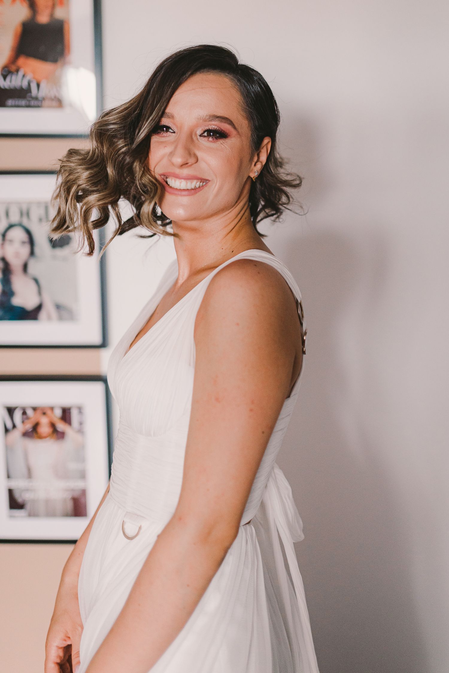 Woman in white sleeveless dress posing against light interior wall with framed artwork.