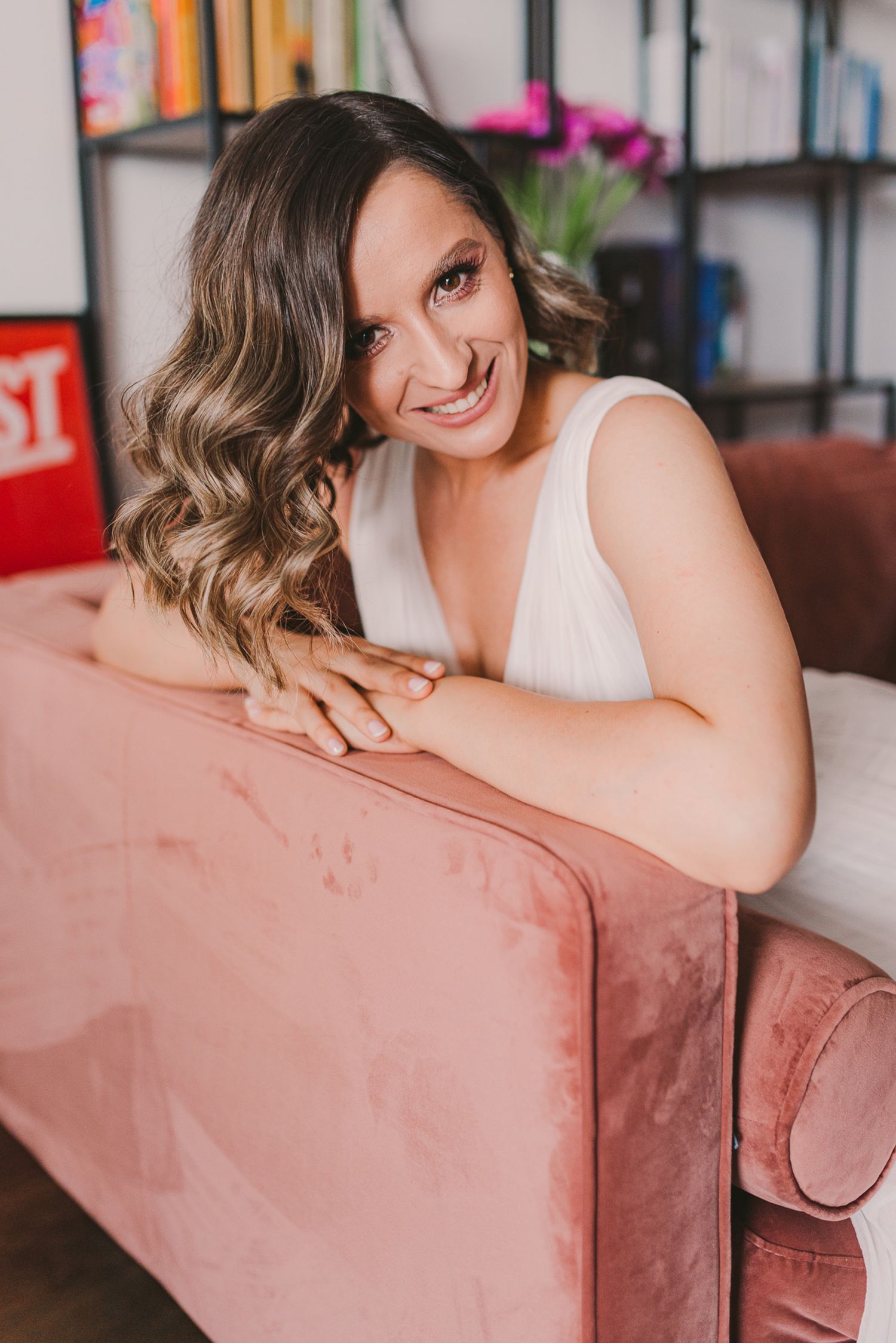 Woman in white dress lounging on pink velvet couch with bookshelves in background.