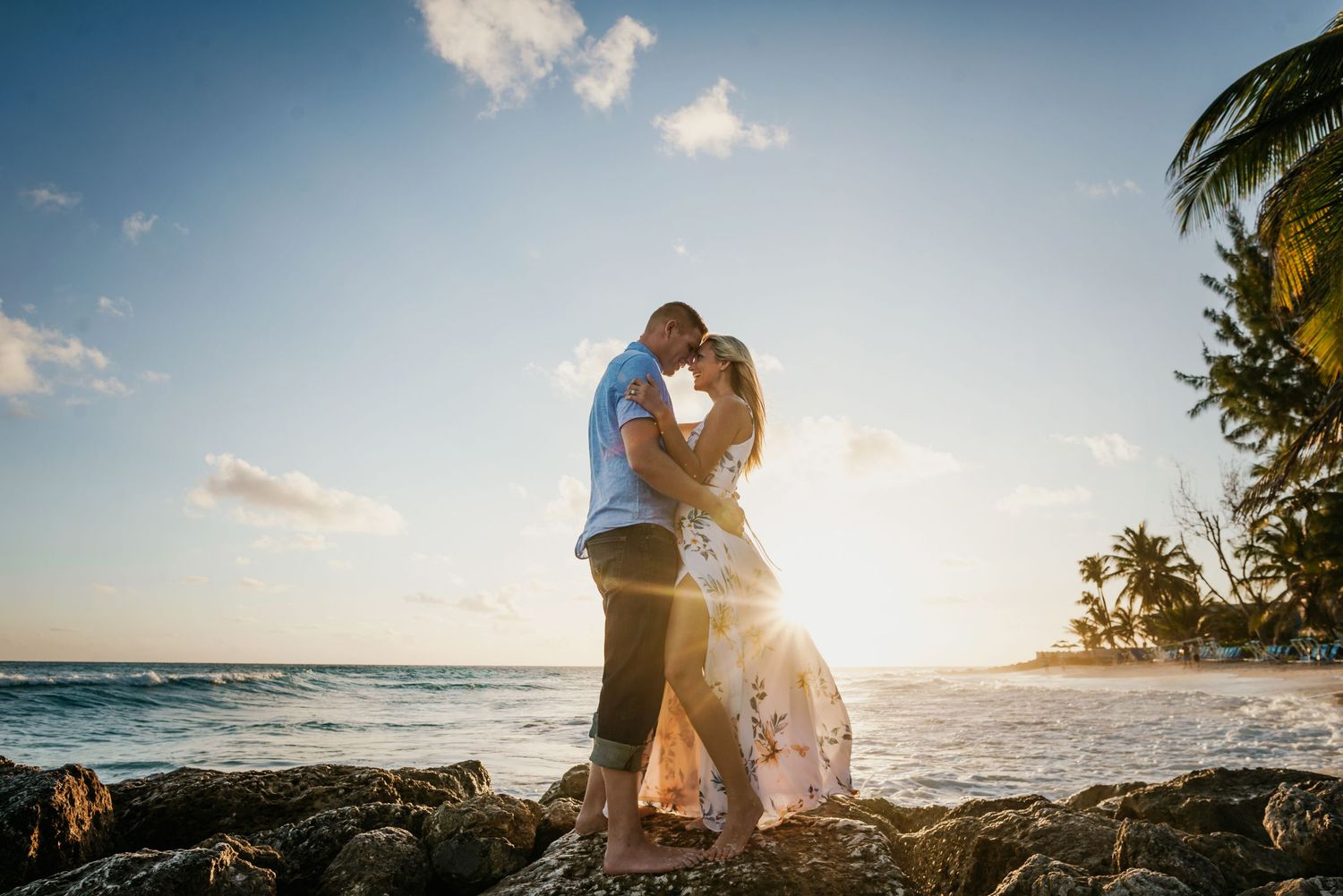 Couple embracing on rocky beach at sunset with palm trees and ocean waves.